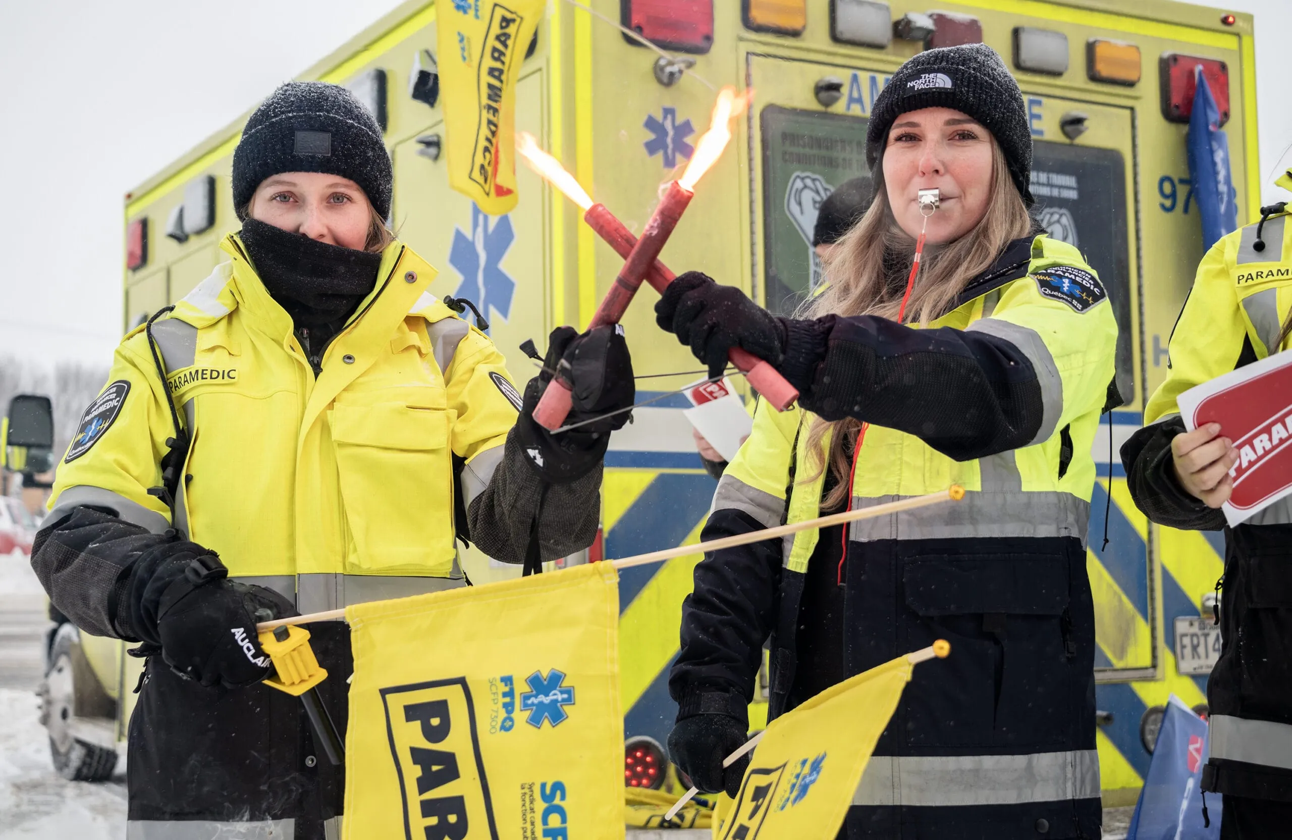 Deux paramédics devant une ambulance, l’une tenant des feux de détresse allumés, l’autre un sifflet à la bouche.